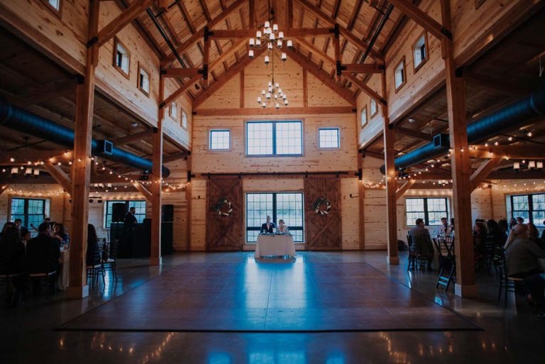 A spacious, rustic barn interior with wooden beams and chandeliers creates the perfect wedding ambiance. A couple sits at a table in the center, under soft lighting. Guests are seated at tables along the sides, while the dance floor remains clear for celebration.