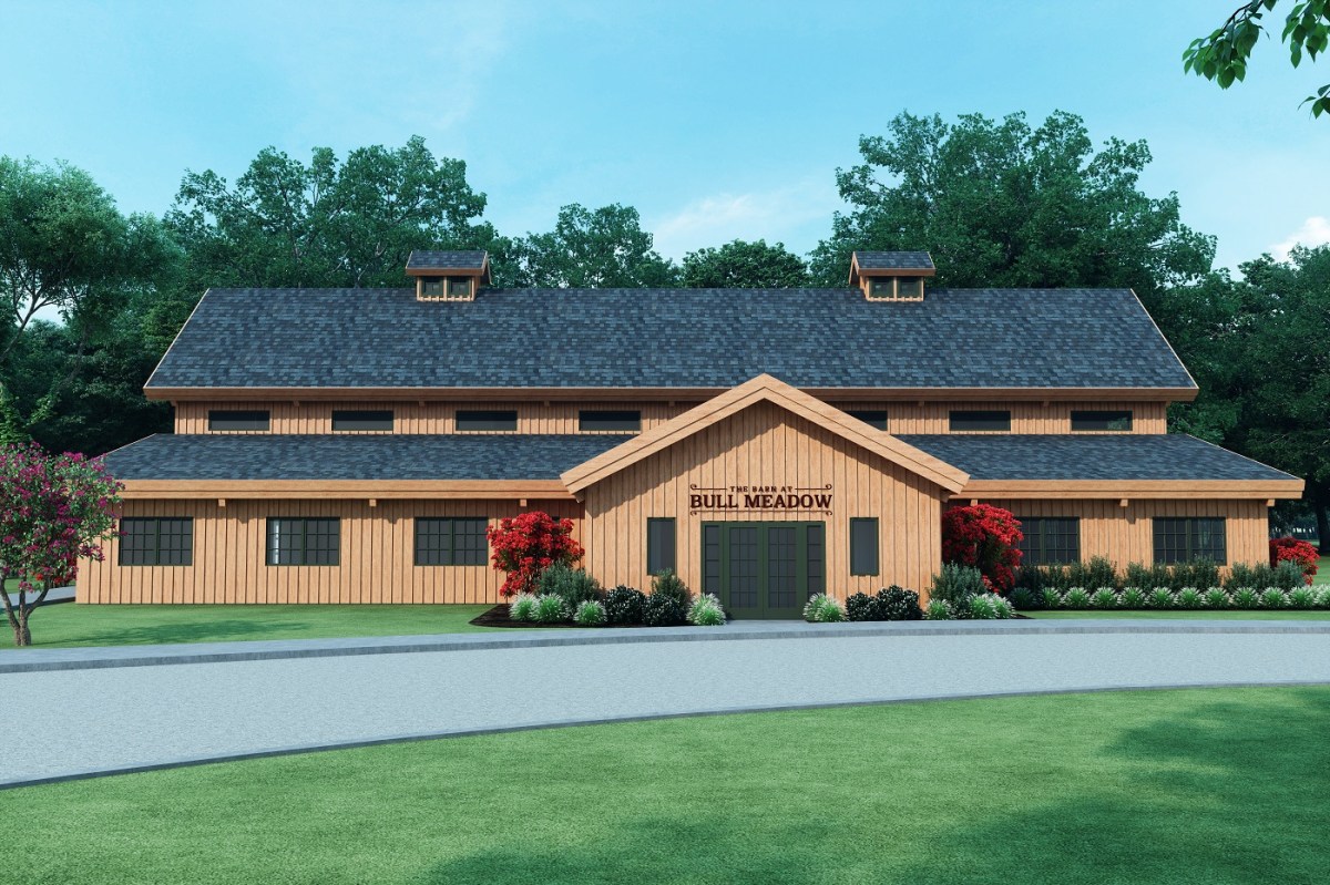 A large, barn-like building with a wooden facade, labeled "Bull Meadow," offers a charming wedding venue. It features a dark shingle roof and is surrounded by lush greenery and landscaped bushes with red flowers. A paved driveway elegantly curves in front, welcoming guests.