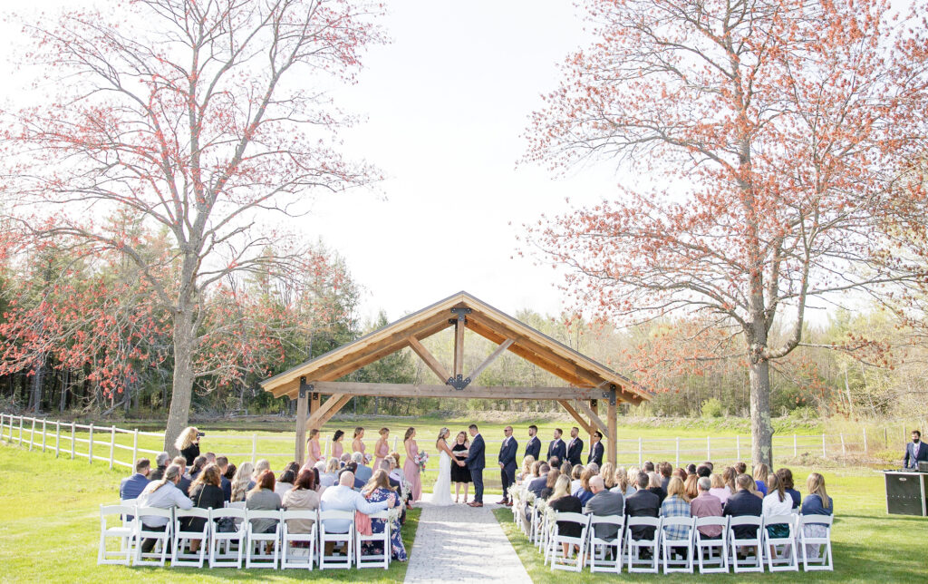 An enchanting wedding unfolds under a wooden gazebo, where the bride and groom stand with the officiant and bridesmaids. Guests sit on white chairs, eyes fixed on the couple. The picturesque setting is embraced by a rustic fence and lush trees.