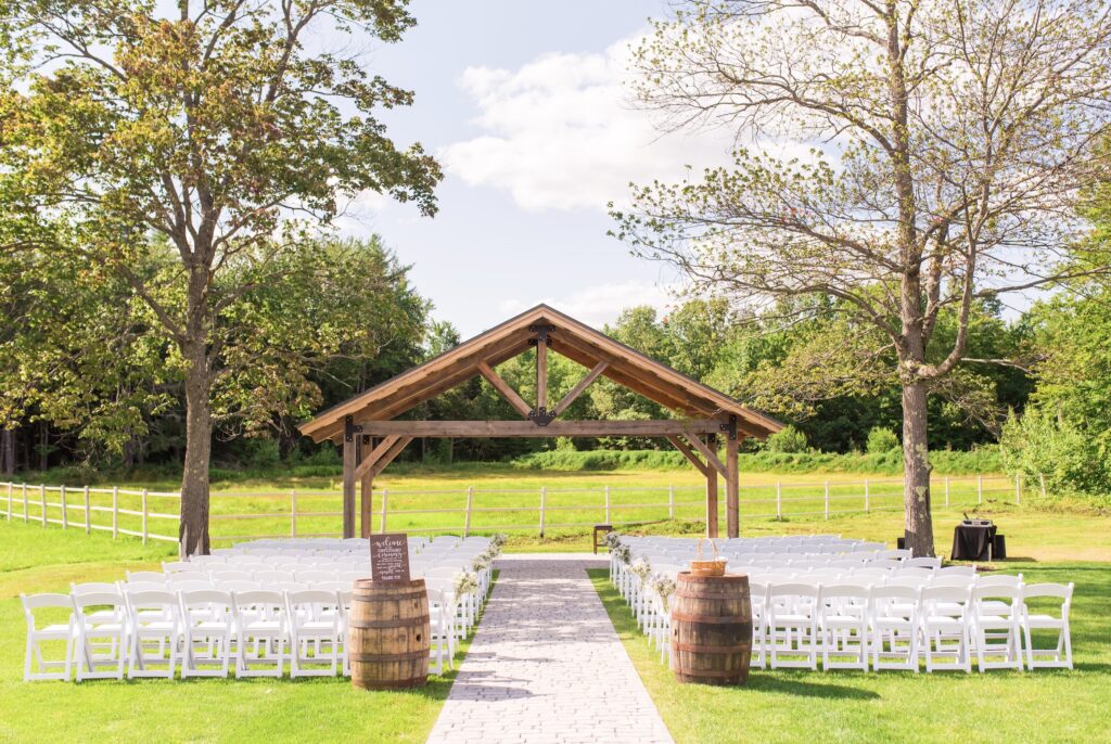 An enchanting outdoor wedding setup features rows of white chairs facing a charming wooden gazebo. Two rustic barrels elegantly flank the aisle, guiding guests toward the gazebo. Lush green grass and towering trees envelop the area under a partly cloudy sky, setting a perfect wedding scene.