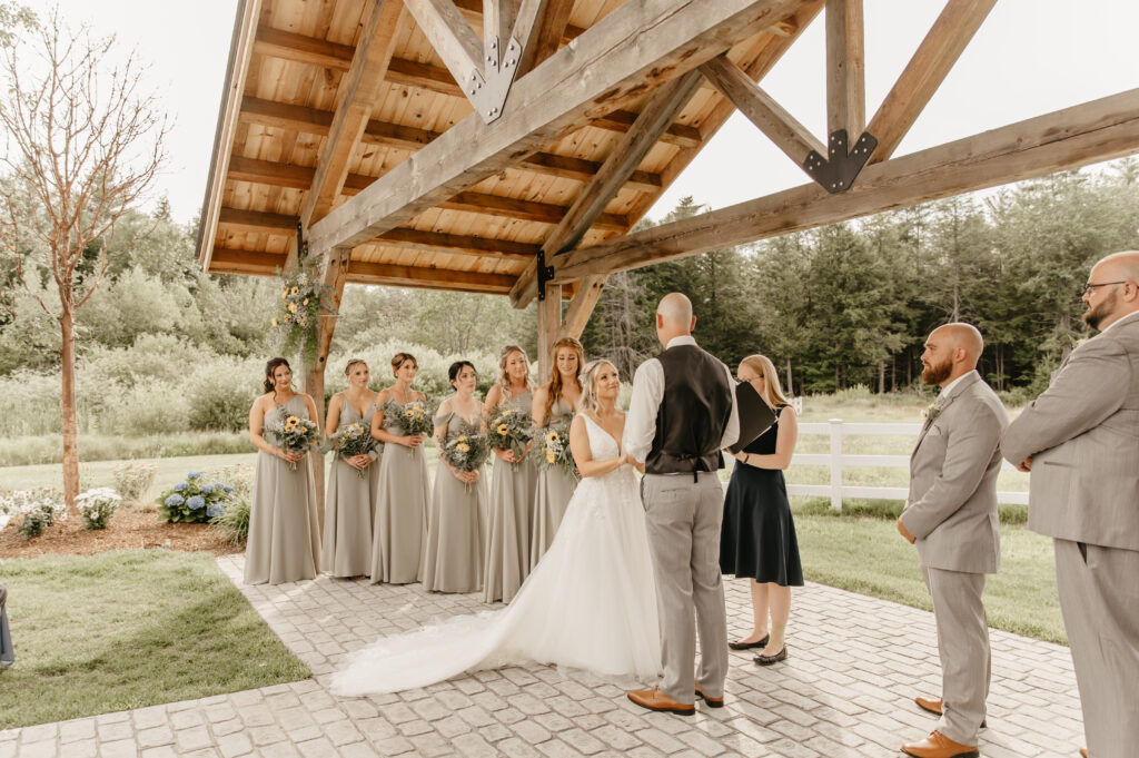 A picturesque wedding unfolds outdoors under a wooden pavilion. The bride and groom stand facing each other, surrounded by bridesmaids in elegant gray dresses and groomsmen in sleek gray suits, set against a backdrop of lush greenery.