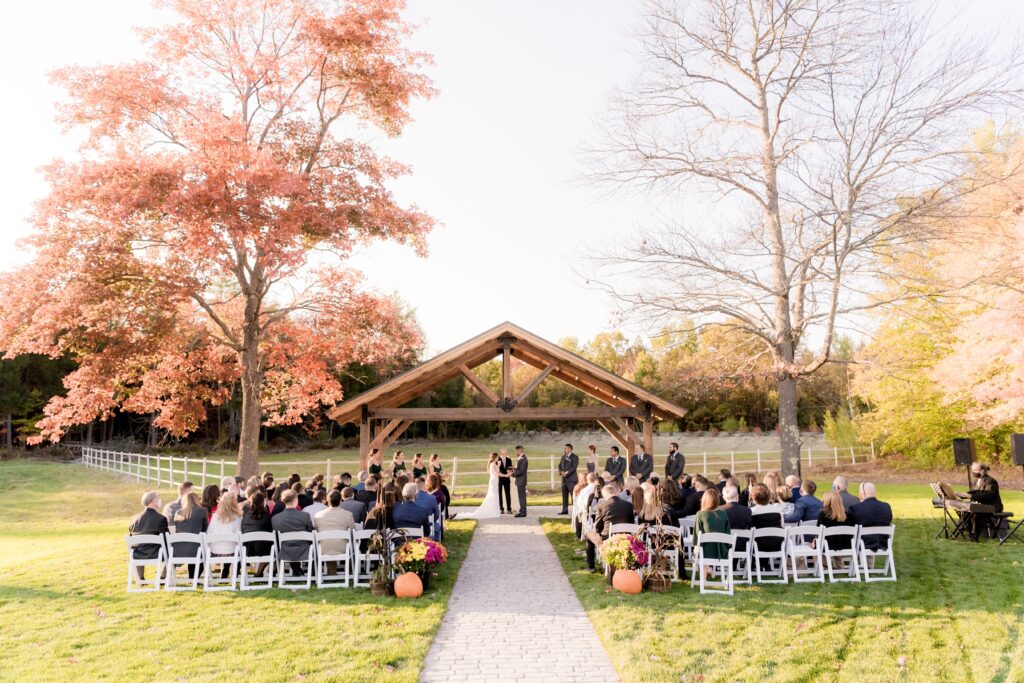 An outdoor wedding ceremony unfolds beneath a charming wooden arbor on a sunny day. Guests are seated on white chairs, facing the couple and officiant. The scene is framed by trees adorned in autumn foliage, with a graceful pathway leading to the heart of this enchanting nuptial celebration.