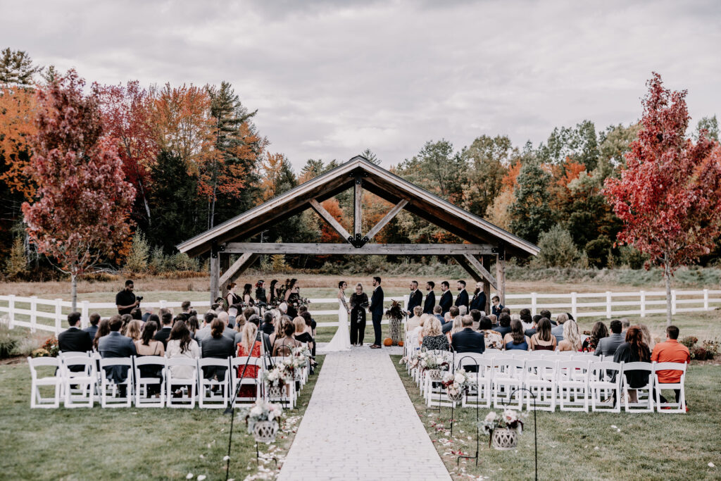 A wedding unfolds at a wooden pavilion, embraced by autumn trees with red and orange leaves. Guests are seated on white chairs along a brick aisle, leading to the couple under the pavilion, as they exchange heartfelt vows.