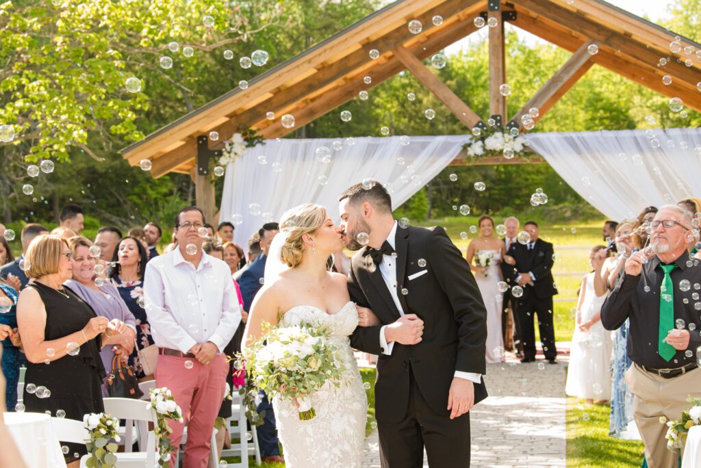 A bride and groom share a wedding kiss under a wooden arch with draped fabric, surrounded by guests. Bubbles float in the sunny outdoor setting, as onlookers smile and clap. The bride holds a bouquet of white flowers and greenery.