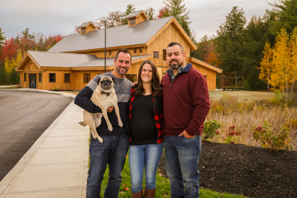 Three people stand smiling in front of a charming, wooden building surrounded by trees. The person on the left holds a pug, and they're dressed in casual, warm attire. It's an autumn wedding scene, with colorful foliage enhancing the joyful atmosphere.