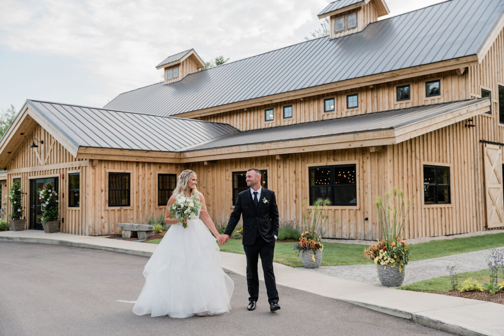 The wedding scene unfolds with a bride in a white gown and a groom in a black suit walking hand in hand before a rustic wooden barn. Smiling at each other, they admire the barn adorned with flowers in large pots against the backdrop of a partly cloudy sky.