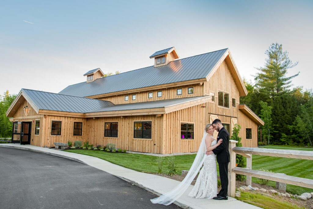 A couple in wedding attire embraces on a pathway beside a rustic wooden barn, setting the perfect scene for their wedding day. The bride wears a long veil and the groom is suited up. Surrounded by lush green lawn and trees under a clear blue sky, their love shines brightly.