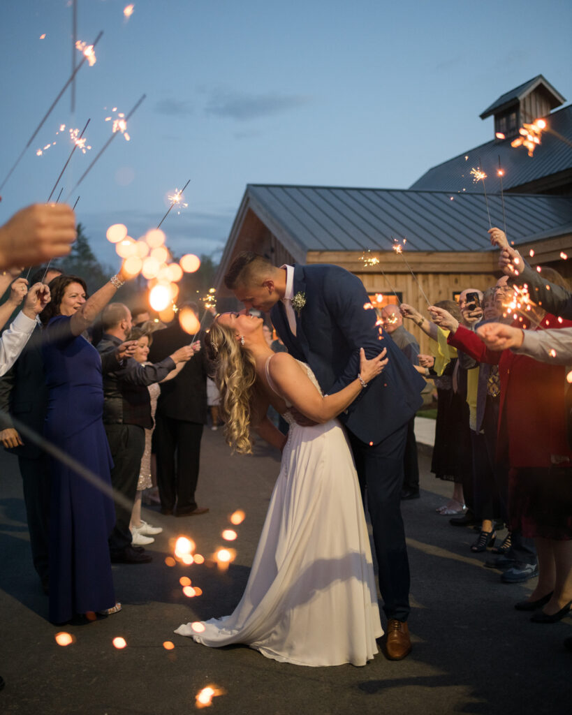 A couple shares a romantic kiss during an enchanting evening wedding, surrounded by guests holding sparklers. The bride wears a white dress, and the groom is in a blue suit. This festive celebration unfolds under the dim evening sky, capturing the joyful essence of their special day.