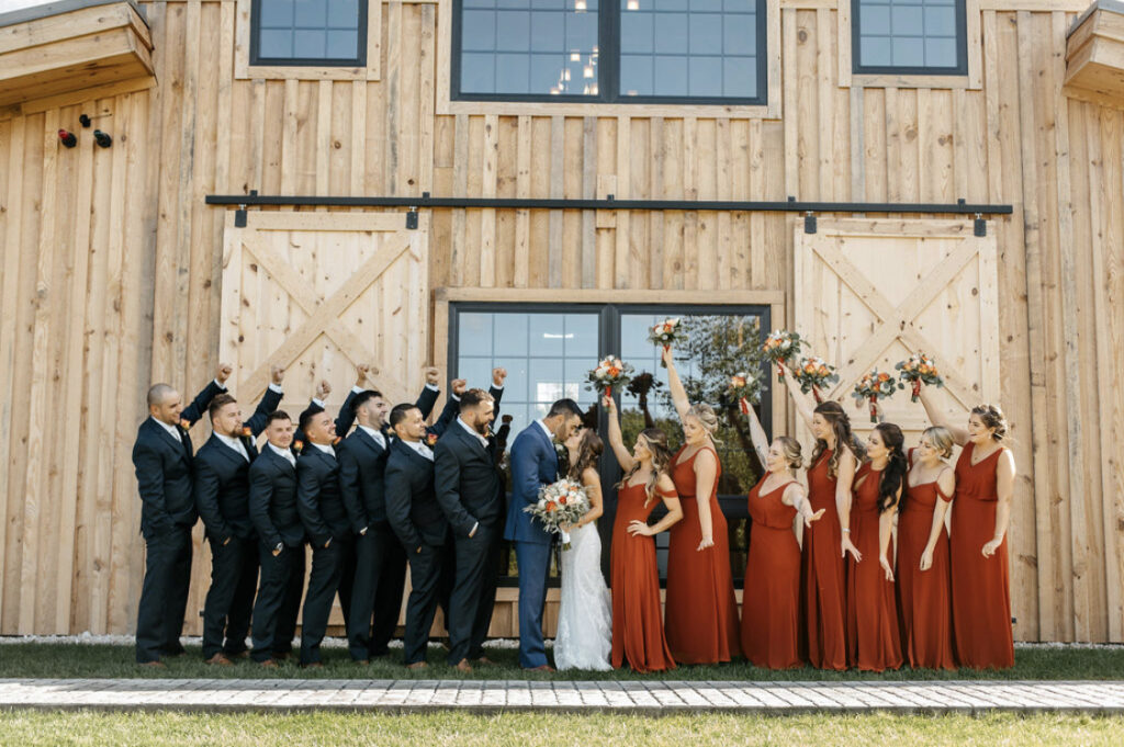 A wedding party stands outside a rustic barn. The bride and groom share a romantic kiss in the center, surrounded by bridesmaids in vibrant red dresses and groomsmen in sharp dark suits, as the bridesmaids jubilantly hold up their bouquets in celebration of this joyous wedding day.