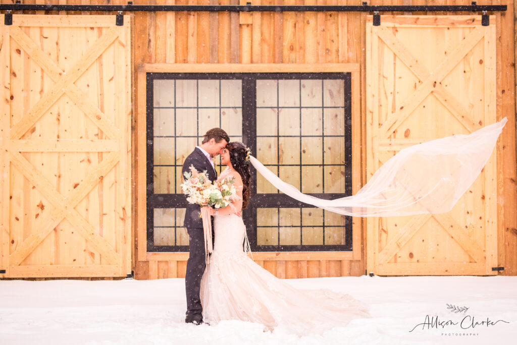 A bride and groom embrace in front of a wooden barn with large doors during their enchanting winter wedding. The bride's veil flows elegantly in the wind, surrounded by snow, crafting a picturesque scene.