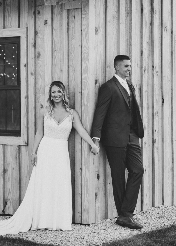 A bride and groom stand smiling in a timeless wedding photo, holding hands on either side of a wooden barn wall. The bride's long white dress contrasts with the groom’s dark suit, captured beautifully in black and white.