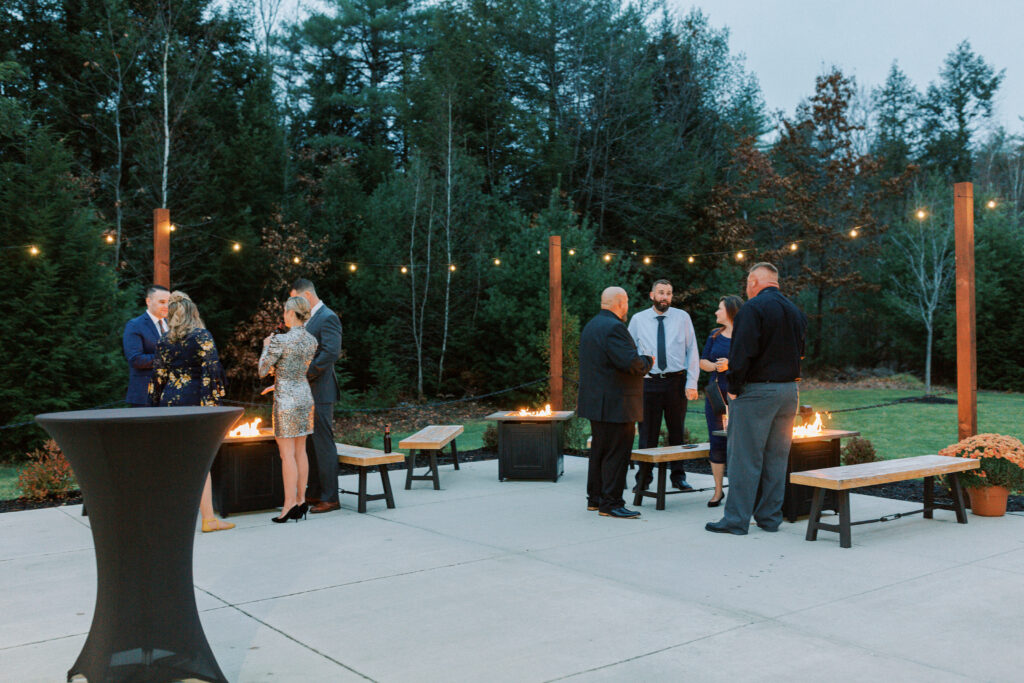 A group of people dressed formally gather and converse outdoors on a patio, celebrating a wedding. String lights glow above, with small fires burning in fire pits amidst the seating. The area is surrounded by trees in the evening light.