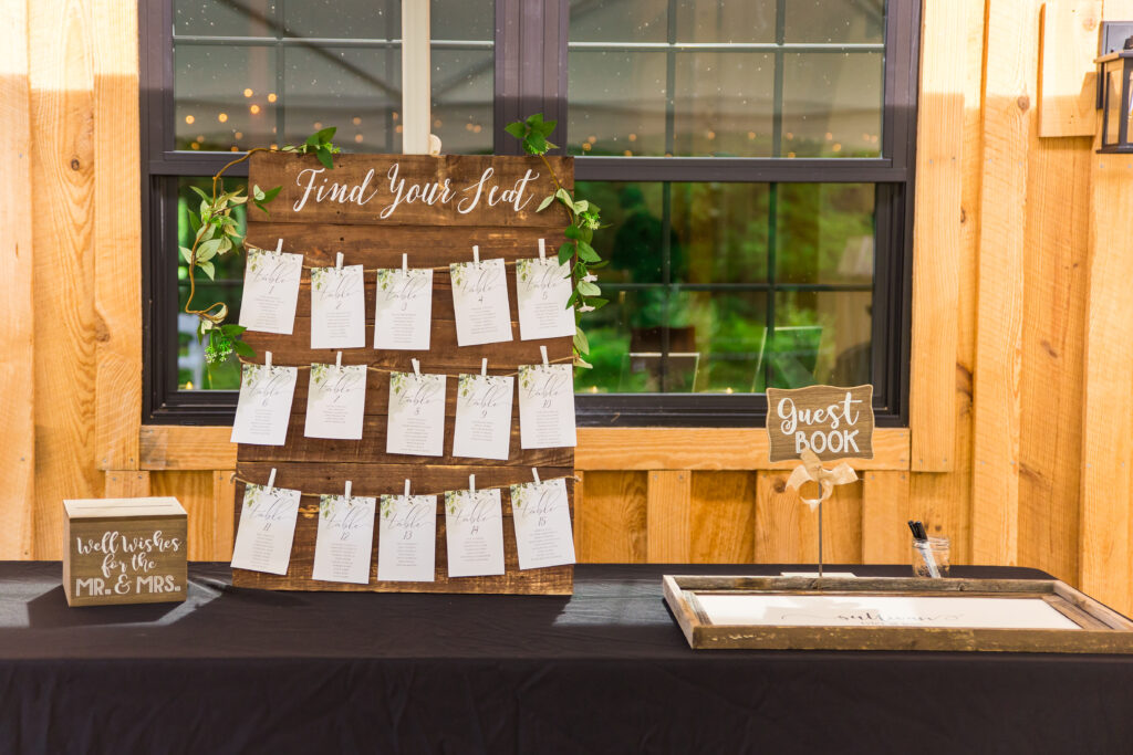 A rustic wedding seating chart graces a wooden board with cards clipped beneath the title "Find Your Seat." Nearby, a guest book on a stand and a box labeled "Well Wishes for the Mr. & Mrs." rest elegantly on a black tablecloth.