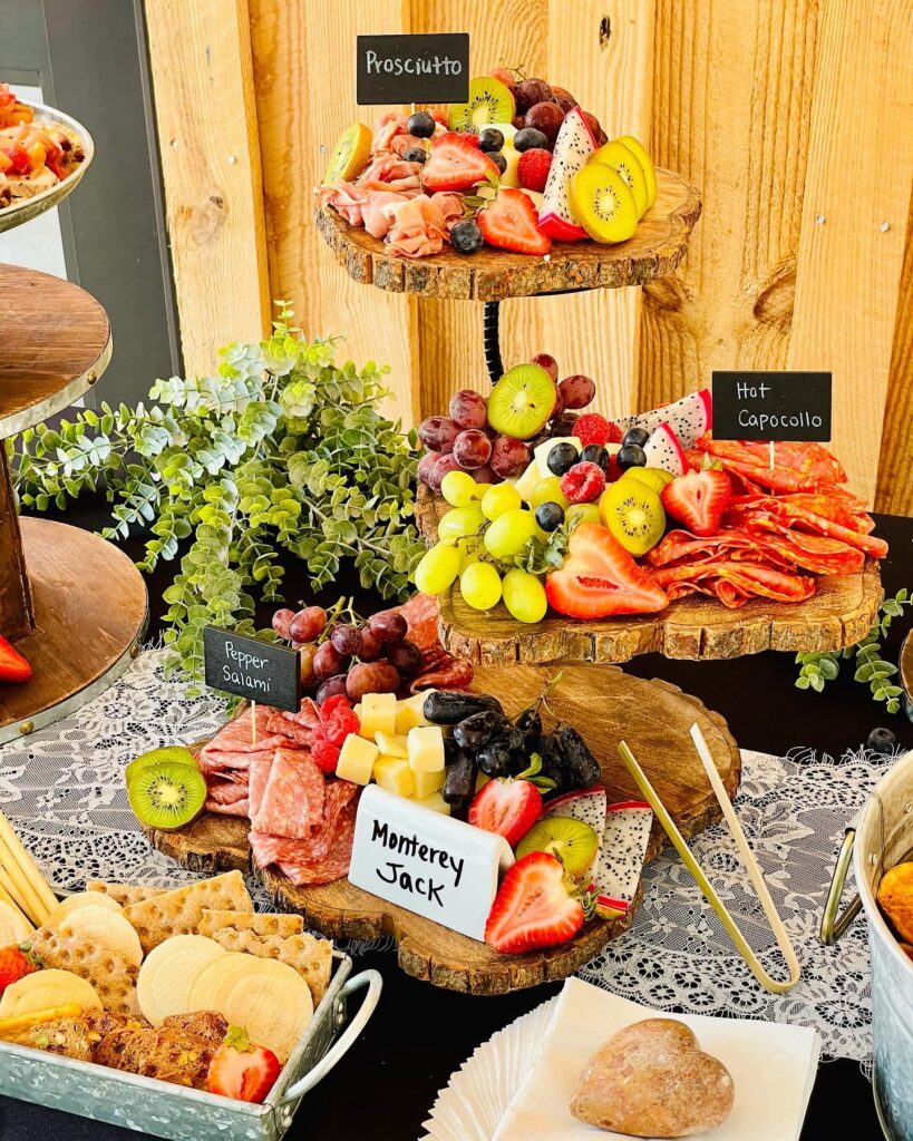 A rustic, wedding-inspired three-tiered charcuterie display features prosciutto, hot capocollo, pepper salami, and Monterey Jack cheese, accented with strawberries and kiwi. Nearby on the lace-covered table lies a small basket of crackers and a roll, perfect for your special day.