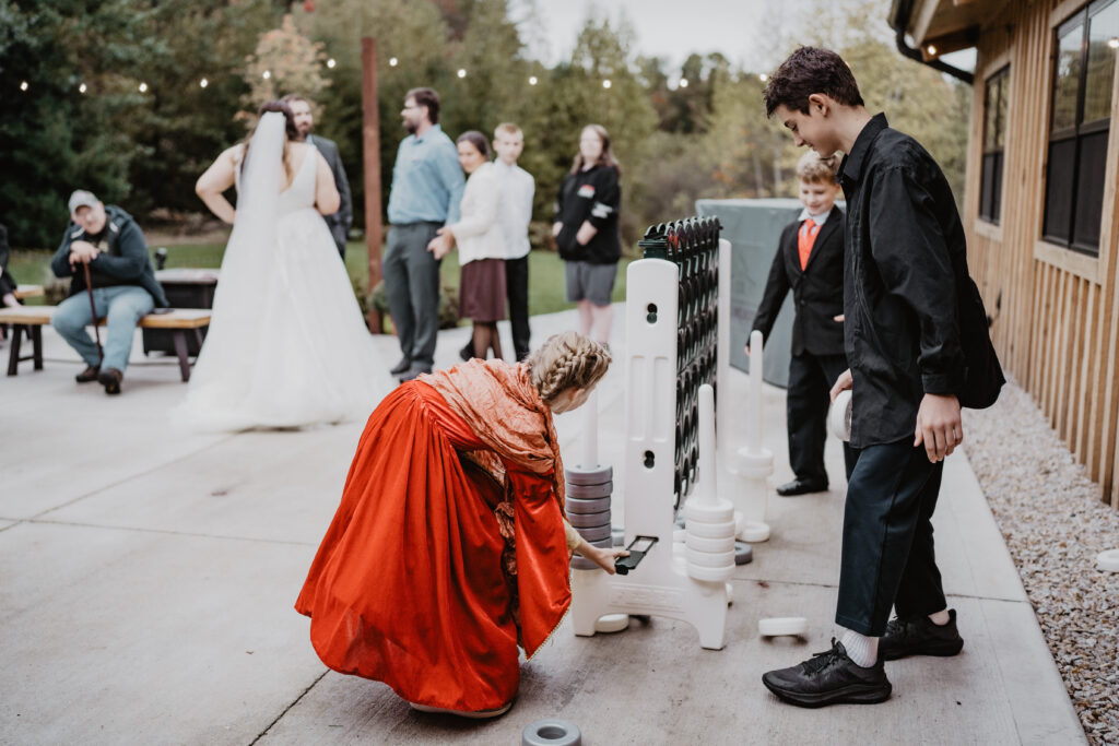 Children play a giant Connect Four game outdoors at a wedding. A girl in a red dress places a piece while a boy in dark clothing watches. Wedding guests and the bride are in the background, with decorative lights twinkling overhead.