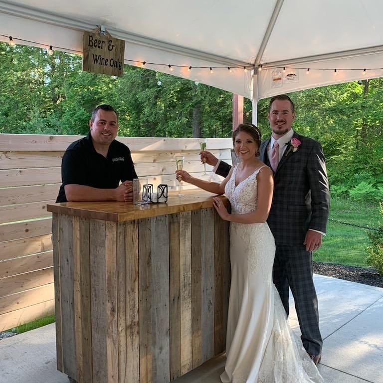 A bride and groom stand at a small rustic wooden bar under a white tent, holding drinks and smiling, perfectly capturing their wedding day bliss. A bartender is behind the bar, providing service. The outdoor setting features green trees in the background, with a sign reading "Beer & Wine Only.