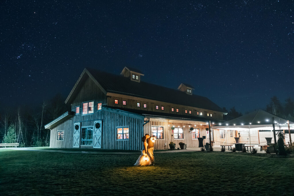 A lit barn stands under a starry night sky, suggesting a romantic wedding setting with its warm glow from inside. A sculpture illuminated in the foreground adds charm, while a canopy adorned with lights hints at an outdoor celebration. Trees grace the background, completing this enchanting scene.