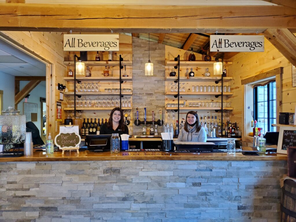 Two people stand behind a wooden bar with a stone facade, reminiscent of an intimate wedding venue. Shelves stocked with various bottles and glassware line the background. Signs above read "All Beverages", though the "Bar Closed" sign rests on the counter. Warm lighting creates a cozy atmosphere.