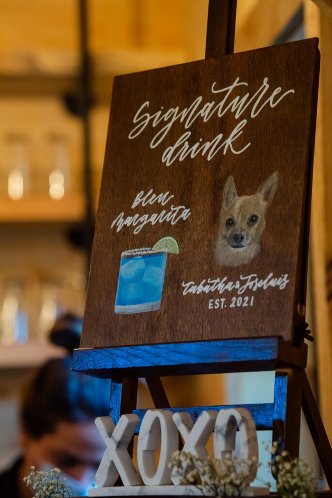 A charming wooden sign reads "Signature Drink" with a blue margarita illustration next to a dog's portrait. Perfect for your wedding, the text features "Est. 2021" and delicate "XOXO" letters at the bottom, all set against a backdrop of blurred shelves.