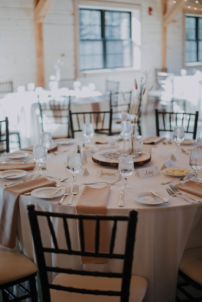 Elegantly set round table with a white tablecloth, glassware, and beige napkins in a sunlit room perfect for a wedding. A wooden centerpiece holds a simple floral arrangement. Black chairs encircle the table, while a window in the background bathes the scene in natural light.