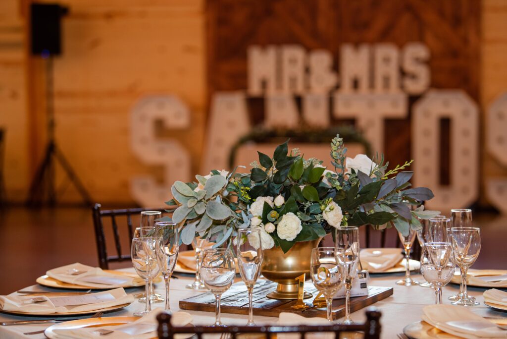 A beautifully set wedding table with an elegant floral centerpiece, surrounded by neatly arranged plates, glassware, and cutlery. In the background, large illuminated letters spell "MR & MRS SANTOS" against a wooden backdrop, capturing the essence of this enchanting wedding celebration.