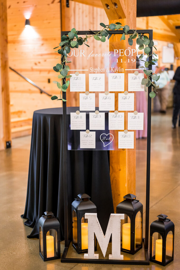 A wedding seating chart elegantly presented on a glass board adorned with lush greenery. It showcases numbered tables and names, standing gracefully on a polished floor beside a black table. Three candle-lit lanterns and a large letter "M" complete this stunning matrimonial display.