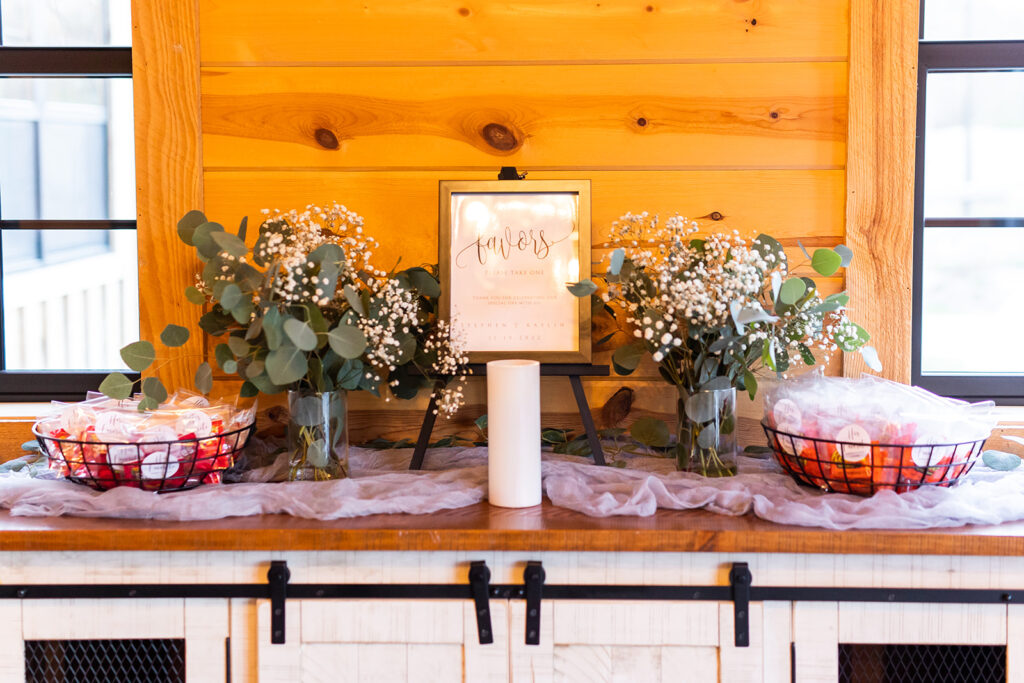 A rustic wedding table display features a "favors" sign surrounded by glass vases with green leaves and white baby's breath flowers. Two black baskets hold small gift bags. The setup is against a wooden wall with black-framed windows on each side.