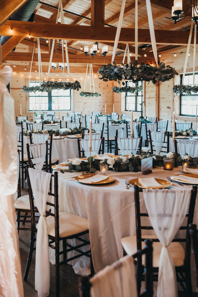 A beautifully decorated wedding reception hall exudes elegance with its round tables set in white tablecloths, elegant place settings, and lush greenery. Wooden beams and hanging wreaths of eucalyptus add rustic charm, while Chiavari chairs surround this enchanting wedding scene.