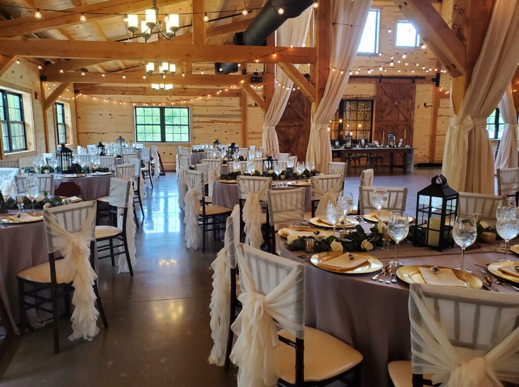 A rustic wedding reception venue with wooden beams and draped curtains. Round tables are elegantly set with plates, glasses, and lantern centerpieces. Soft lighting and a polished floor enhance the cozy wedding ambiance.