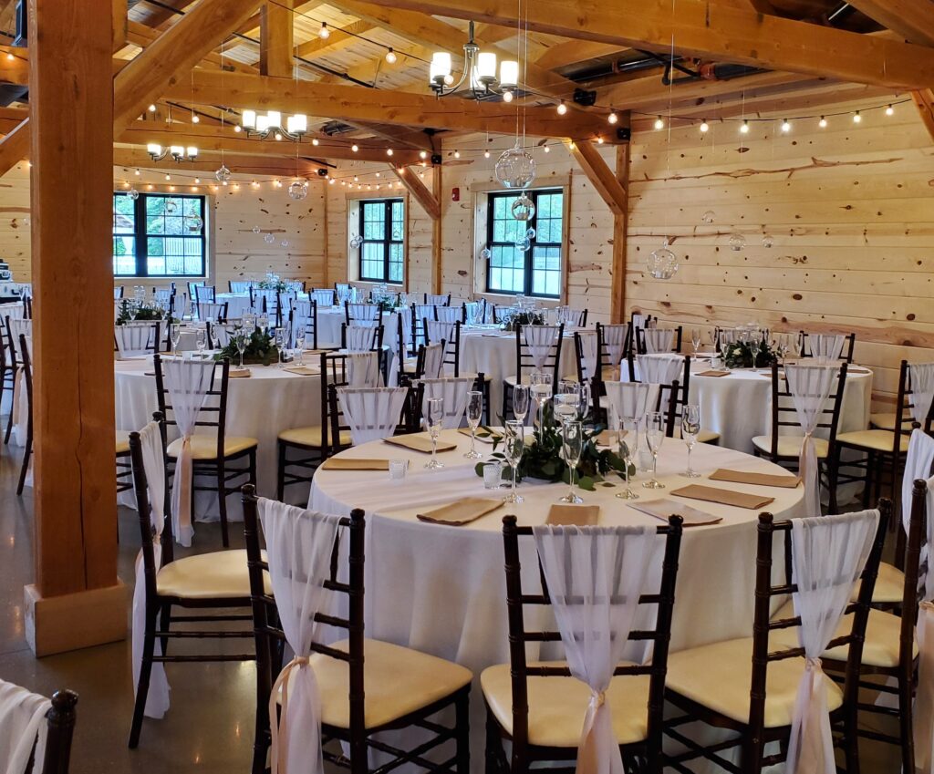 A beautifully decorated reception hall with wooden walls and beams sets the perfect wedding scene. Round tables, covered in white tablecloths, are adorned with green and white floral centerpieces. Elegant chairs with white fabric accents surround the tables, creating a romantic atmosphere.