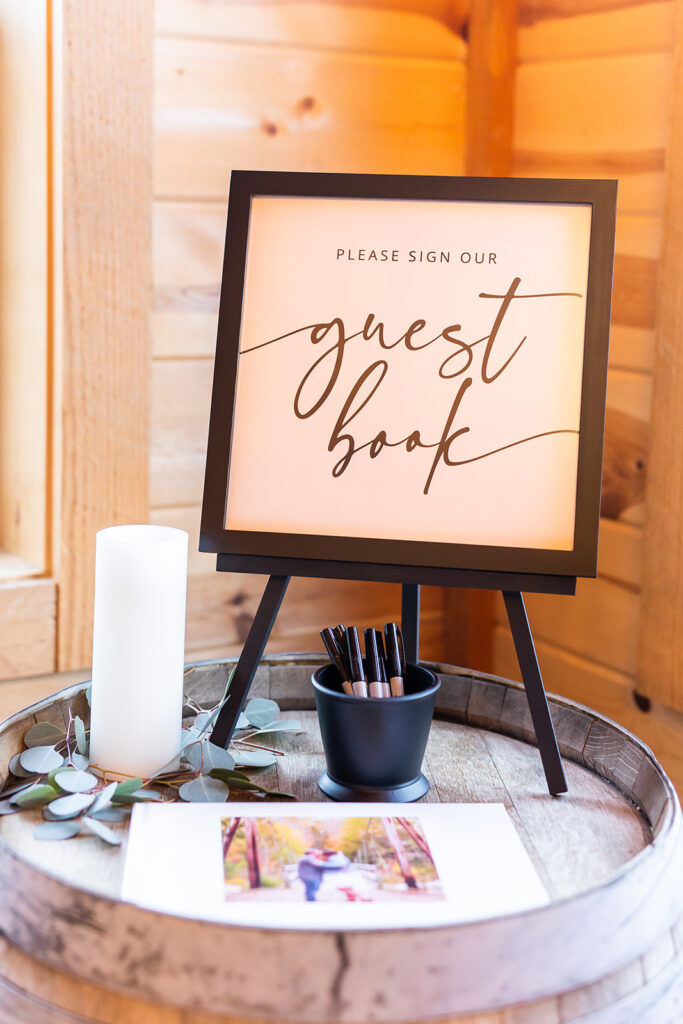 A wooden barrel serves as a table, holding a glowing sign that reads, "Please sign our wedding guest book" in elegant script. A photo album and a small black pot of pens are nearby, illuminated by a candle and decorated with green leaves.