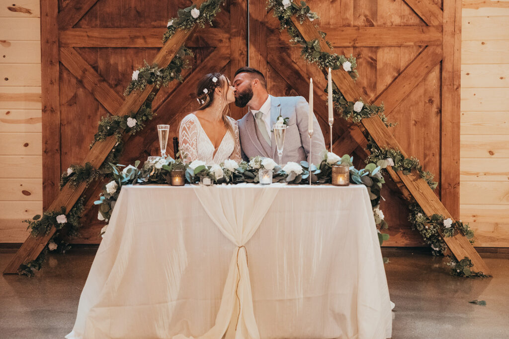 At their enchanting wedding reception, the bride and groom share a kiss at a beautifully decorated table, lush with foliage and candles. Behind them, rustic wooden doors are adorned with greenery, creating a perfect romantic backdrop for their special moment.