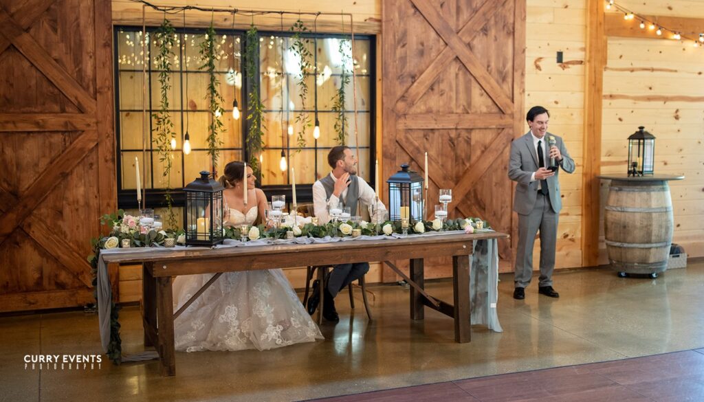 A bride and groom sit at a rustic wooden table adorned with floral and candle decorations, capturing the essence of a charming wedding. Nearby, a man in a suit gives a heartfelt speech in the warmly decorated venue, complete with wooden accents and twinkling string lights.