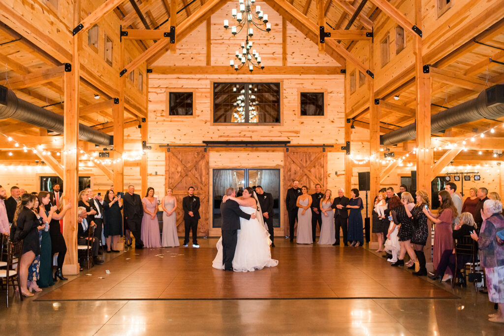 A couple shares their first wedding dance in a rustic wooden barn venue with guests gathered around. Warm lighting and chandeliers enhance the cozy atmosphere. Bridesmaids in matching dresses stand nearby, and the room is filled with joyful onlookers.