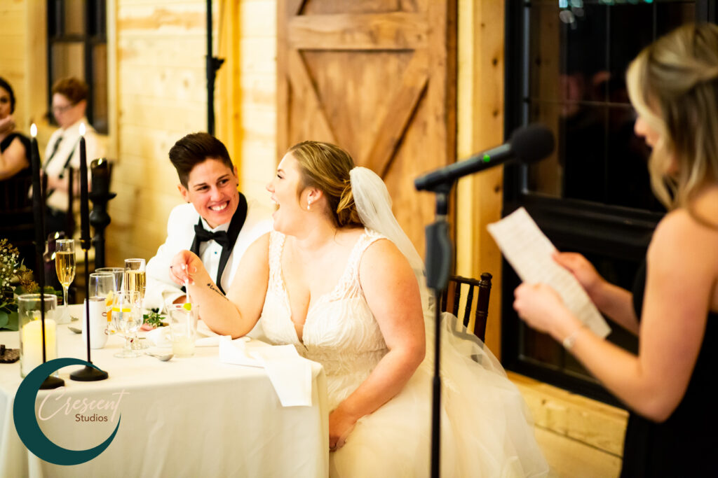 A couple in wedding attire sits at a reception table, smiling as they listen to a heartfelt wedding speech. The bride wears a white gown and veil, while the other person dons a tuxedo. A microphone and speaker are in the foreground, with elegant table settings surrounding them.