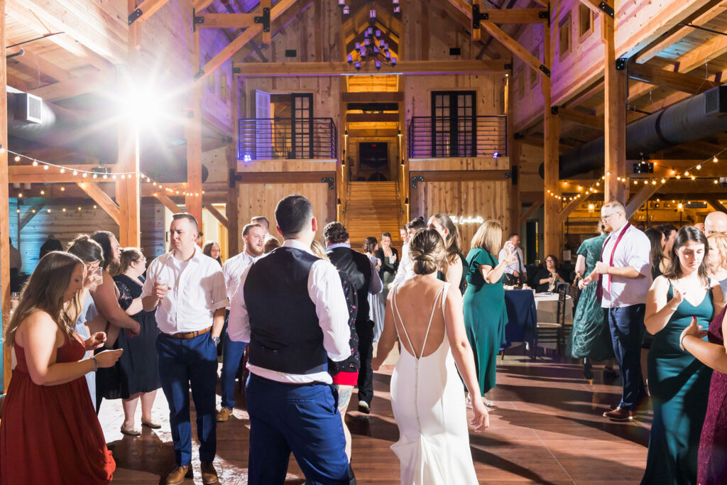 A lively wedding reception unfolds in a rustic wooden venue where guests, dressed in formal attire, dance energetically under the glow of string lights. The bride, elegantly clad in white, faces away from the camera as joy fills the air.