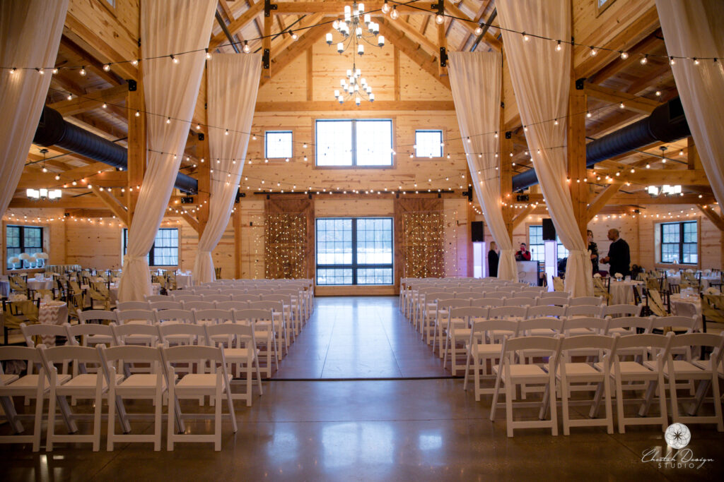 A rustic wedding venue with wooden beams and high ceilings, adorned with string lights and white curtains. Rows of white chairs await the ceremony, facing a twinkling backdrop. Guests gather near the back, ready to witness the beautiful wedding unfold in this enchanting setting.