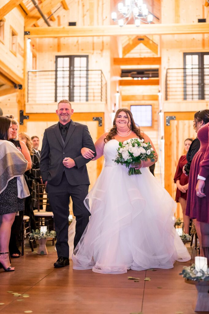 A bride in a flowing white gown walks down the aisle at her wedding, holding a bouquet of white flowers. She is accompanied by a man in a dark suit. They are in a wooden, rustic venue with guests seated on either side.