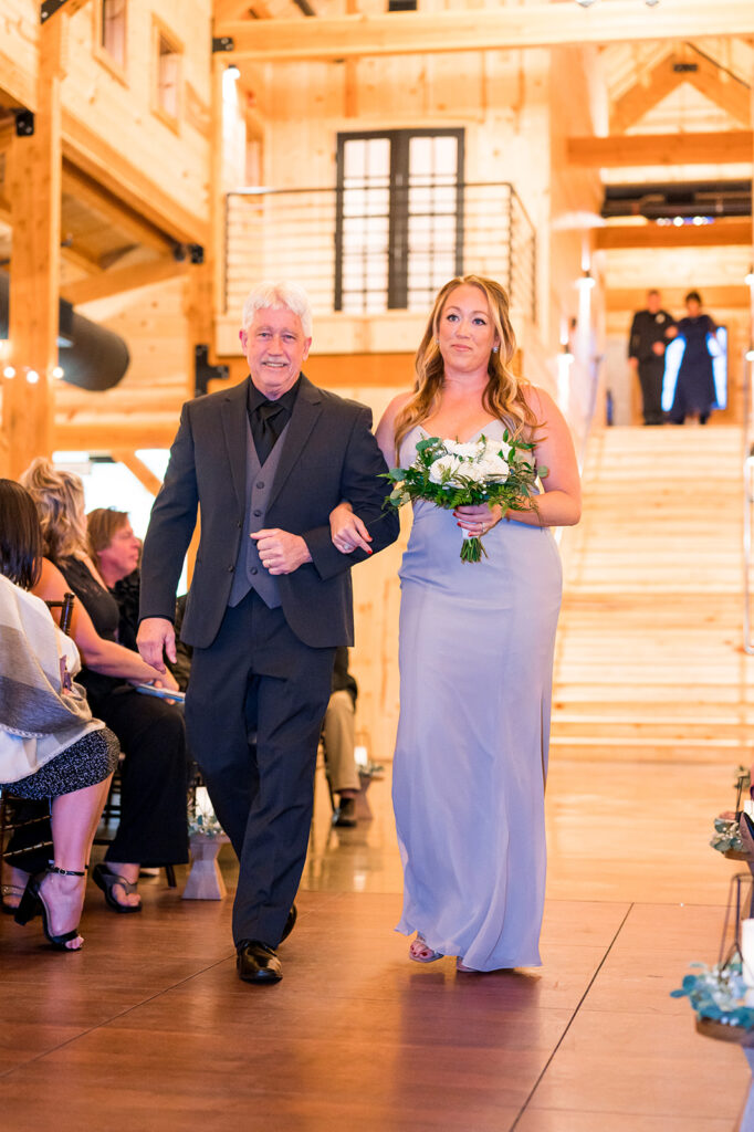 A man in a black suit and a woman in a lavender dress, holding a bouquet, walk arm-in-arm down the wedding aisle in a warmly lit wooden venue. Guests are seated on either side, and a staircase is visible in the background.