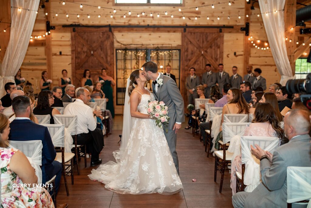 A couple shares a tender kiss at the end of a wooden aisle in a rustic wedding venue, surrounded by guests on either side. The bride holds a bouquet of pink flowers, and wedding string lights hang above, casting a warm, intimate glow over the cherished moment.