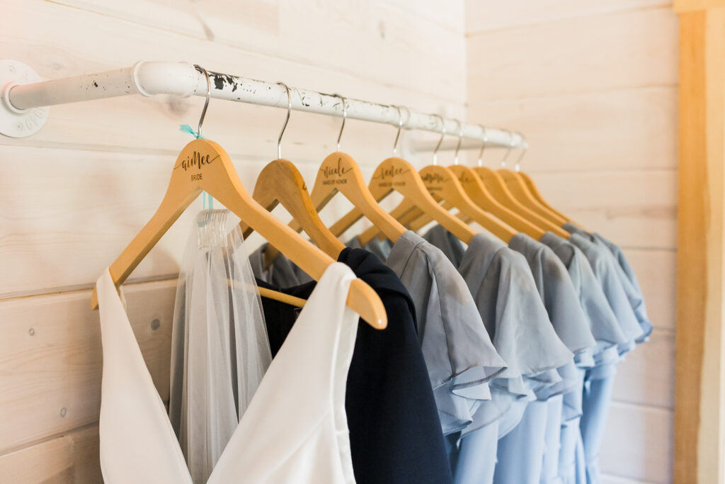 A row of wooden hangers, each engraved with names, holds a selection of dresses in elegant shades of white and blue on a sleek metal rod against a light wooden wall. Perfect for a wedding, the collection features a stunning white bridal gown alongside several blue bridesmaid dresses.