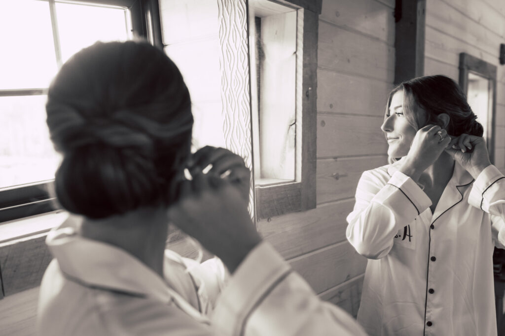 Two people in monogrammed pajamas adjust their earrings, preparing for a special day, as they look out the window of a rustic room. Captured in black and white, this serene scene embodies a candid pre-wedding moment.