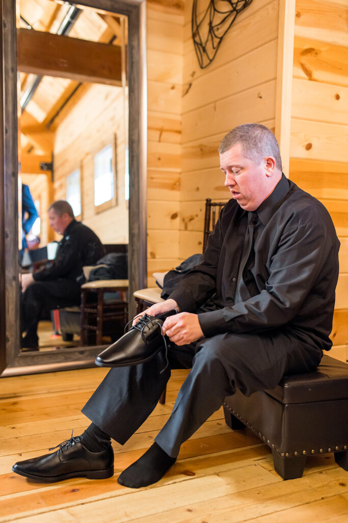 A man in a black outfit sits on a bench in a wooden room, putting on black dress shoes for the wedding. A large mirror reflects his image, showing the room's warm, rustic interior with light wooden walls and floor.