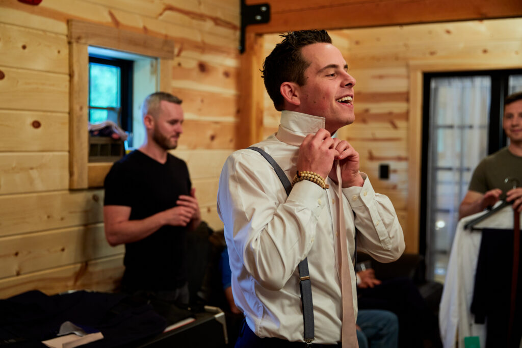 In a rustic, wood-paneled room, a man in a white shirt and suspenders smiles as he adjusts his tie, savoring the joyful pre-wedding moments. Two other men linger in the background, one holding clothes and another standing nearby, enhancing the relaxed atmosphere.