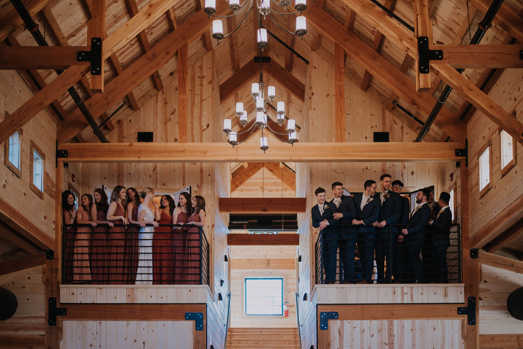 A wedding celebration unfolds as a joyous party stands on two balconies in a rustic wooden venue. Bridesmaids in burgundy dresses grace the left, while groomsmen in dark suits gather on the right. The charming interior boasts exposed beams and twinkling hanging lights, setting a perfect stage for this memorable occasion.