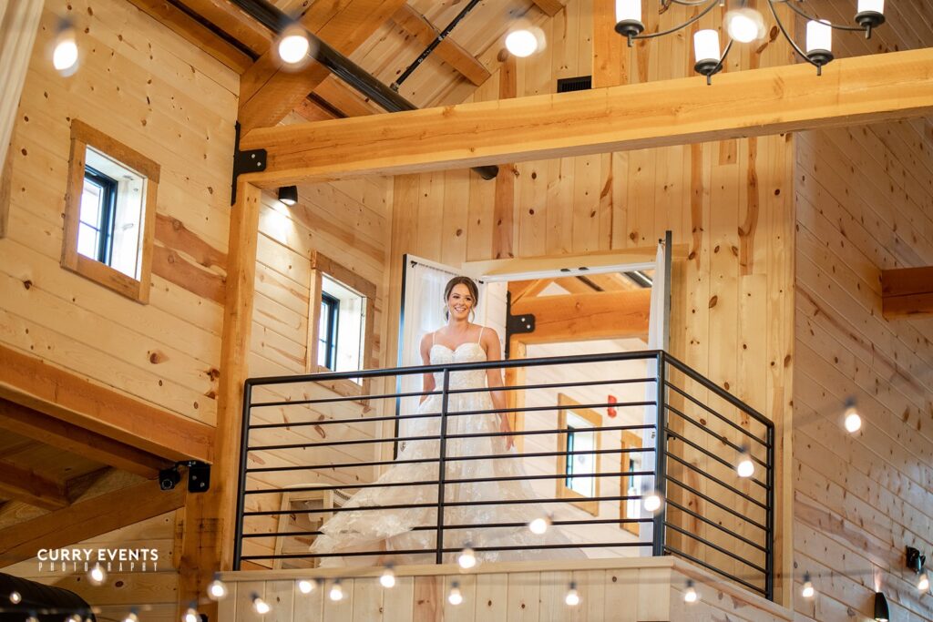 A bride in a white wedding gown stands on a wooden balcony inside a rustic wedding venue. String lights hang from the ceiling, adding a festive touch to the cozy, warmly lit interior.