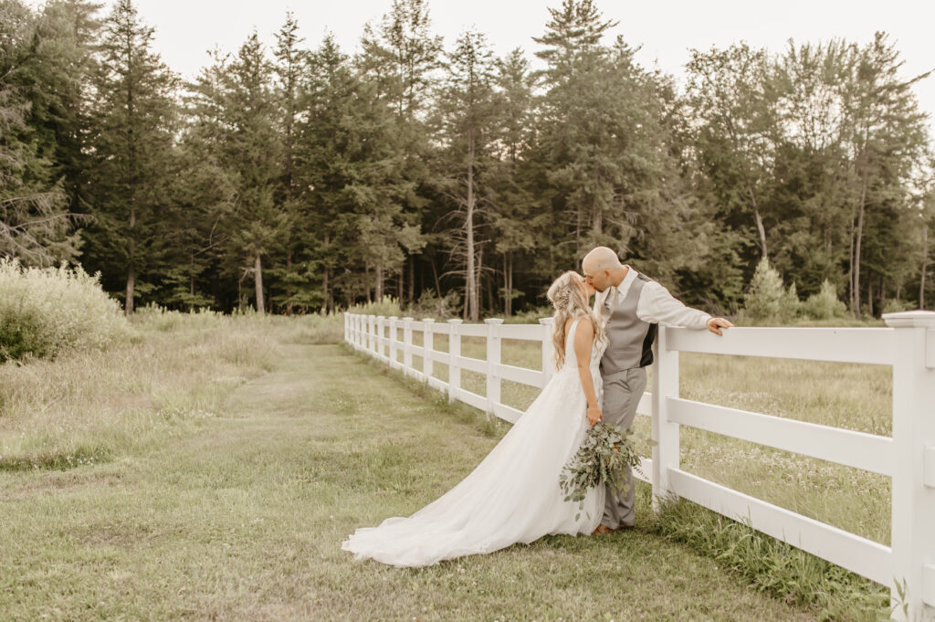 At a charming wedding, a bride and groom share a tender kiss by a white fence in a meadow. The bride, in her long white gown, holds a bouquet, while the groom looks dashing in his vest and tie. Tall trees stand as silent witnesses under the clear sky.