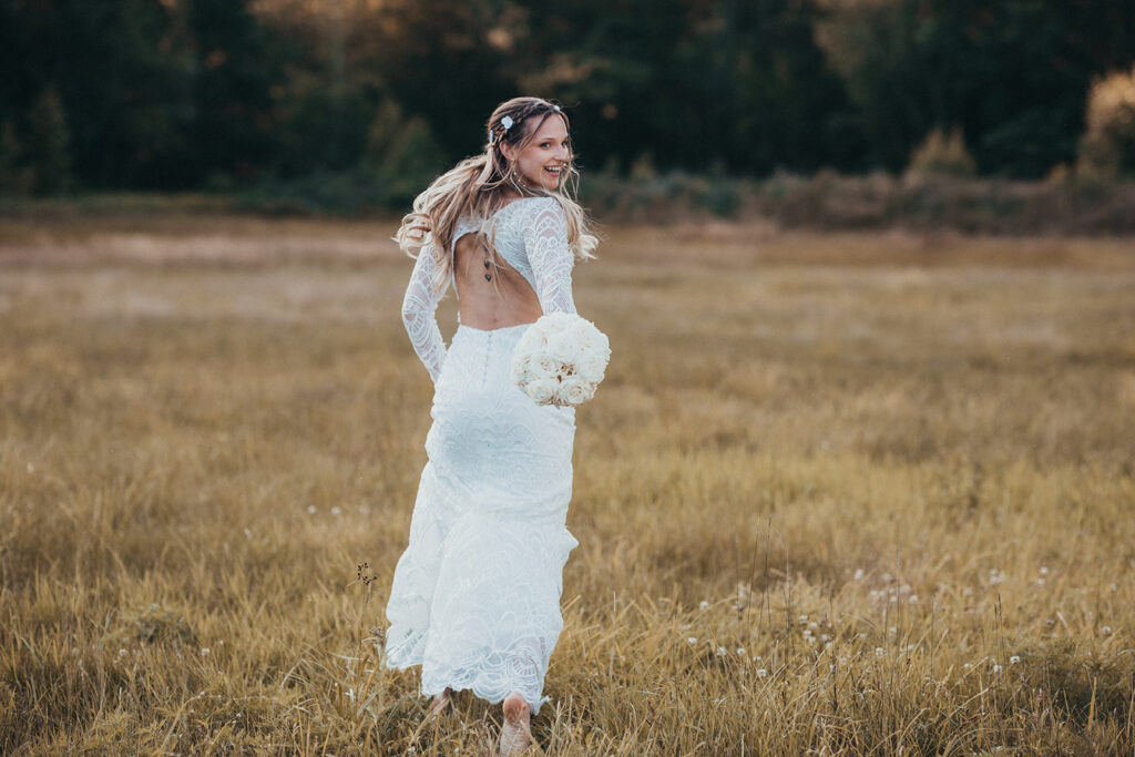 A bride in a long, white lace dress runs joyfully across a grassy field during her wedding, clutching a bouquet of white flowers. The sun sets behind her, casting a warm, golden glow that perfectly captures the magic of the moment.