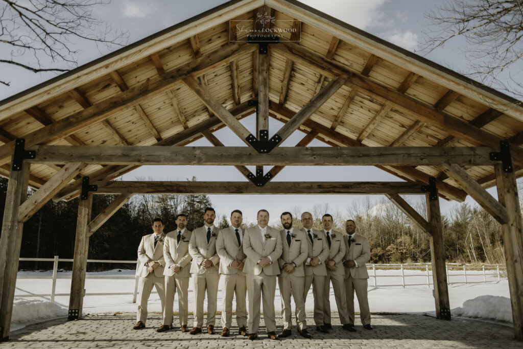 A group of ten men in beige suits stand under a large wooden gazebo, celebrating a winter wedding against the backdrop of a snowy landscape. The sky is clear, and the bare trees hint at the serene chill of the season.