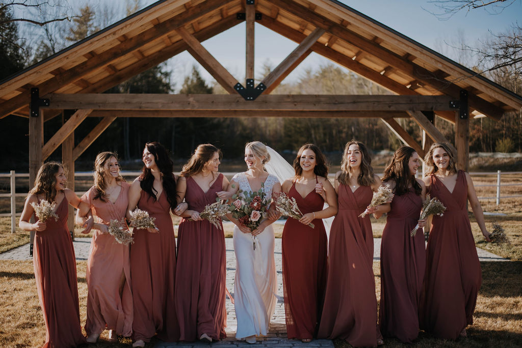 A bride in a white gown and veil stands with her bridesmaids, all dressed in various shades of dusty pink. They are outdoors at a wedding venue, posing in front of a wooden arch and smiling while holding bouquets of flowers.